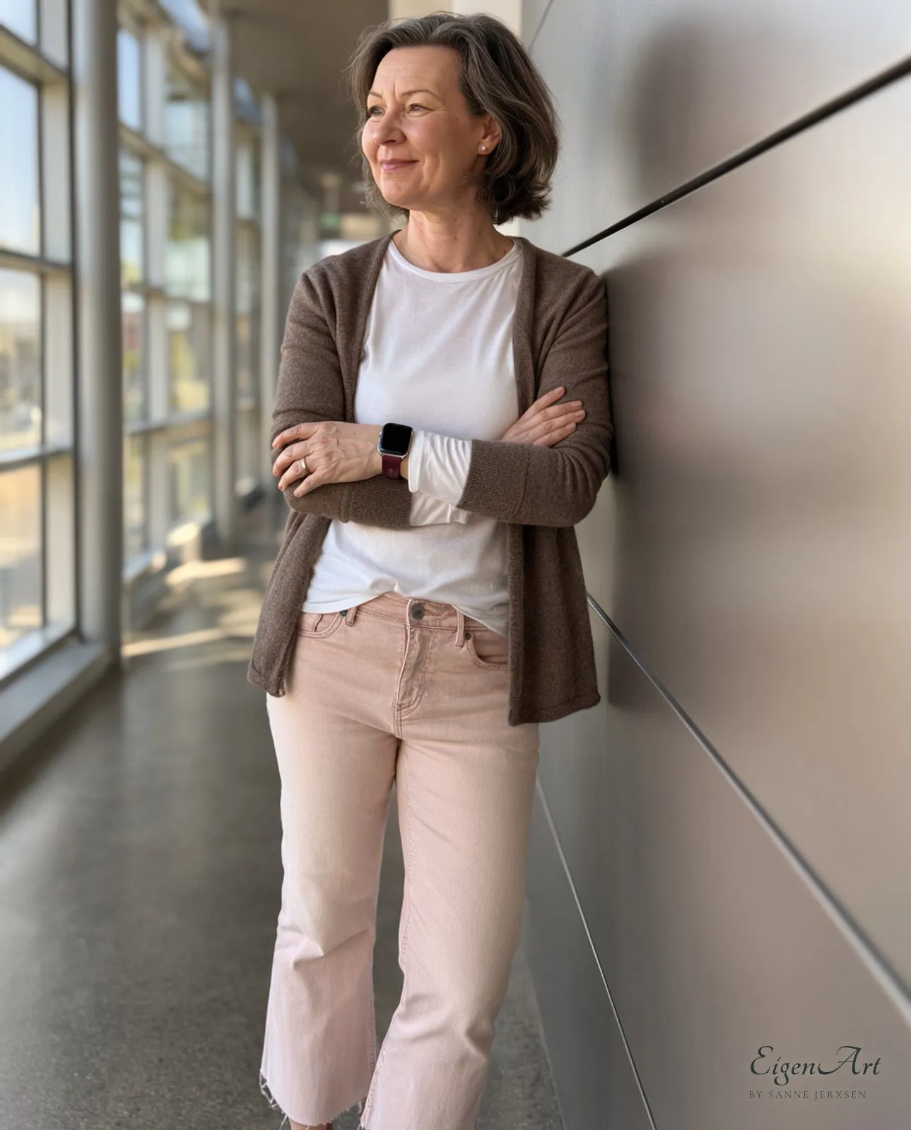 Sanne Jerxsen lehnt mit verschränkten Armen an der Wand eines langen, verglasten Gangs und blickt lächelnd zur Seite, brauner Cardigan, weißes Shirt, hellrosa Jeans